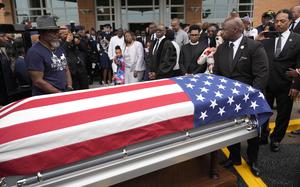 A crowd of people in formal attire watch as a casket draped in the American flag departs.