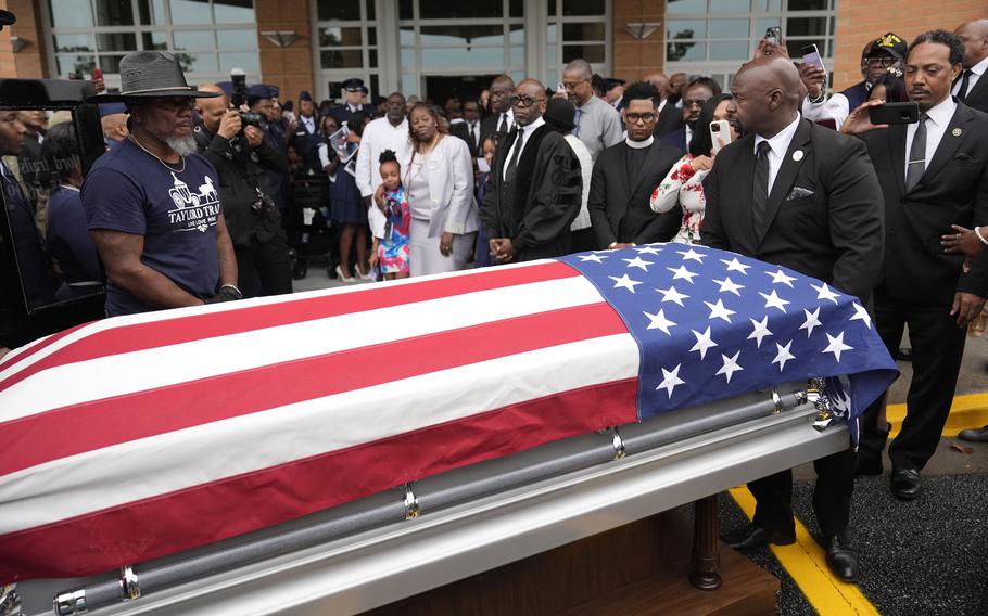 A crowd of people in formal attire watch as a casket draped in the American flag departs.