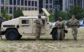 Three soldiers in camouflage uniforms stand next to military vehicle parked on a grassy lot, with city buildings in the background.