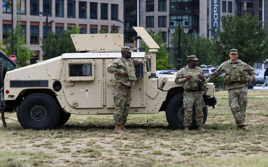 Three soldiers in camouflage uniforms stand next to military vehicle parked on a grassy lot, with city buildings in the background.