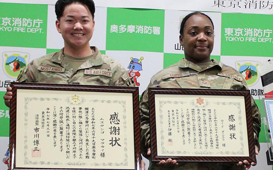 Two U.S. Air Force service members in camouflage uniforms hold framed certificates with Japanese text at a Tokyo Fire Department ceremony. Behind them is a green and white backdrop with Tokyo Fire Department logos and Japanese text. 