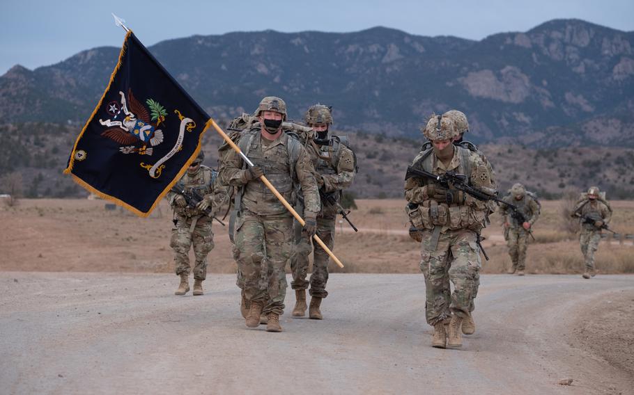 Soldiers in masks march on a road with mountains in the background; one is carrying a flag.