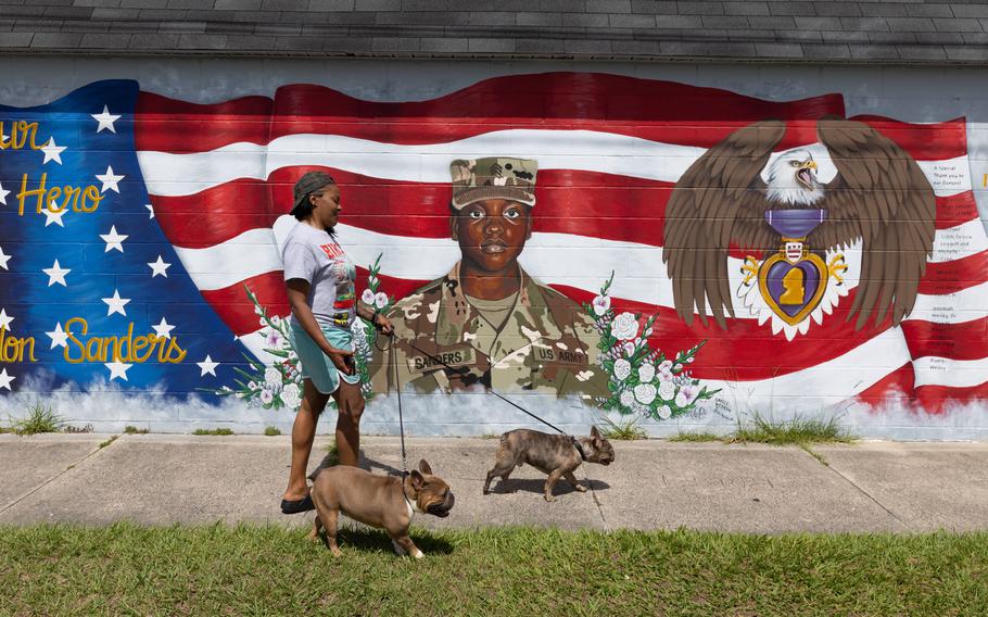 A woman walks two dogs next to a wall with a mural of a fallen soldier.