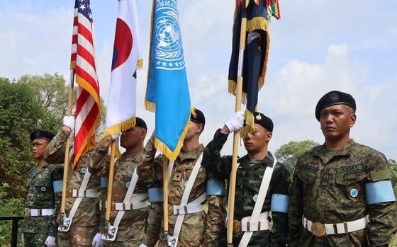 Military honor guard ceremony featuring soldiers in dress uniforms holding flags including the American flag, Japanese flag, UN flag and other national flags. 