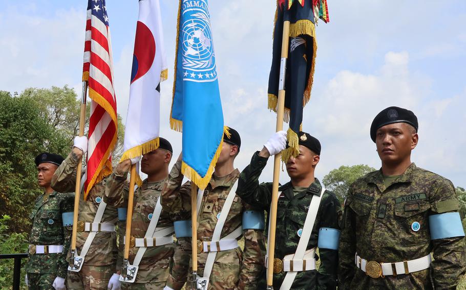 Military honor guard ceremony featuring soldiers in dress uniforms holding flags including the American flag, Japanese flag, UN flag and other national flags. 