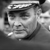 A black and white portrait of a man looking intently at another person whose out-of-focus head takes up the left side of the photo.
