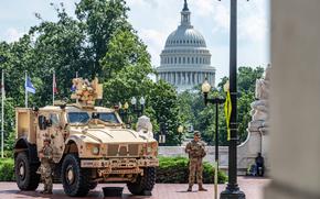 Two soldiers stand alongside a military vehicle with trees and the U.S. Capitol in the background.