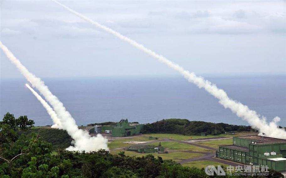 Multiple missiles launch from a coastal military base, leaving white smoke trails as they ascend over the ocean. The installation features green buildings surrounded by hills and vegetation. A CNA watermark is visible in corner.