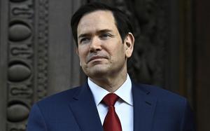 Close-up view of Marco Rubio, wearing a dark suit and red tie, standing in front of a government building.