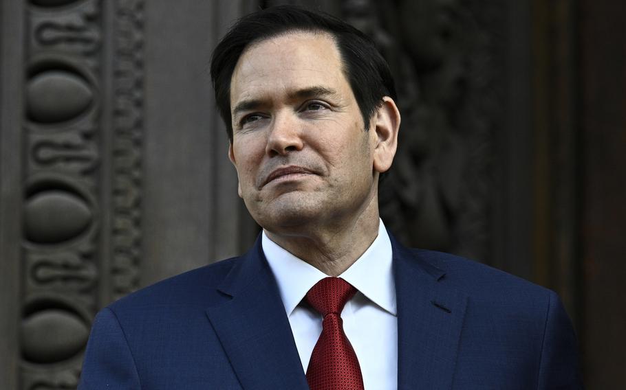 Close-up view of Marco Rubio, wearing a dark suit and red tie, standing in front of a government building.