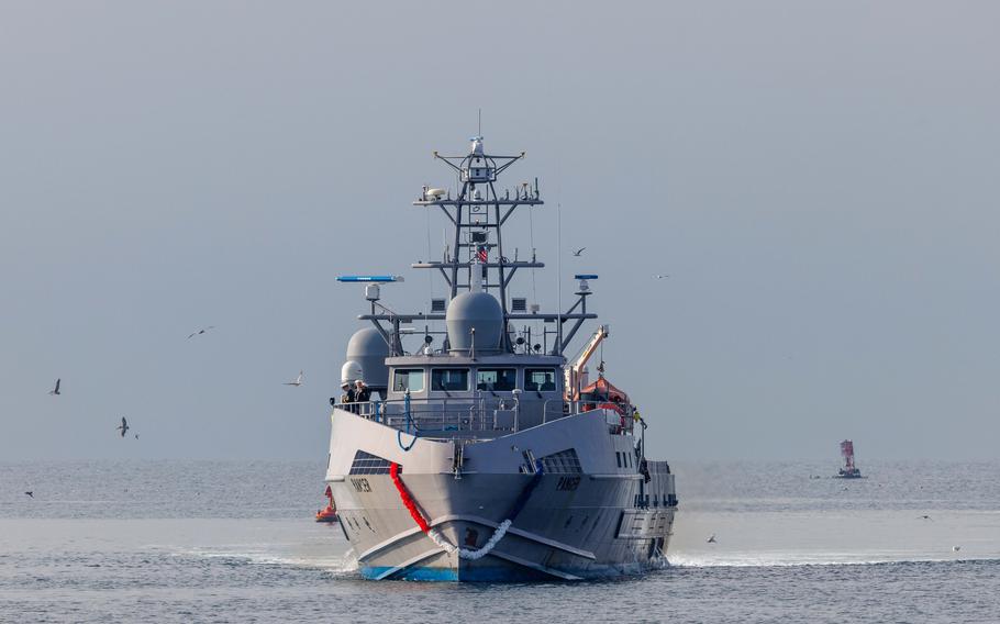 A military patrol vessel approaches through calm waters creating spray at its bow. The gray ship features a tall mast with multiple radar dishes and communication equipment, spherical radar domes and a bridge structure with multiple windows. 