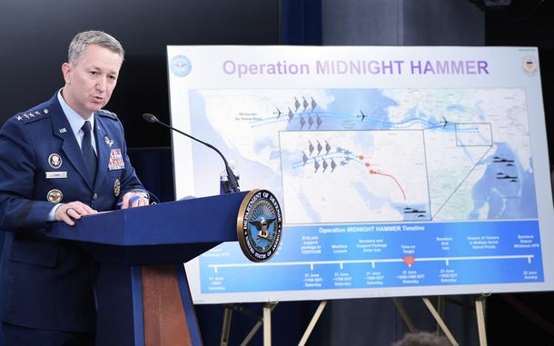 A military officer in blue dress uniform stands at a podium and speaks into a microphone next to an easel holding a large poster with “Operation Midnight Hammer” written at the top above a map of Iran with military movements diagrammed.