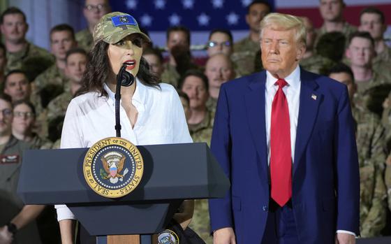 Gretchen Whitmer stands at a podium and speaks into a microphone while Donald Trump stands next to her in front of military members in camouflage uniforms.