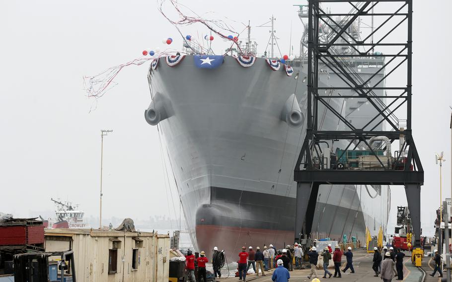 A large gray naval vessel decorated with patriotic bunting and a blue flag with white star sits in a shipyard, surrounded by industrial cranes and construction equipment, with shipyard workers in hard hats gathered below.