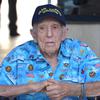 An elderly man is shown seated while wearing a blue shirt with U.S. flags and Navy ships and planes and a hat reading “Pearl Harbor Survivor.”