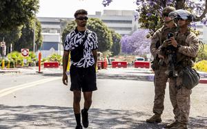 A man wearing a black and white floral shirt and sunglasses walks past two military service members in camouflage combat gear at a checkpoint on a street with a multi-story office building in the background.