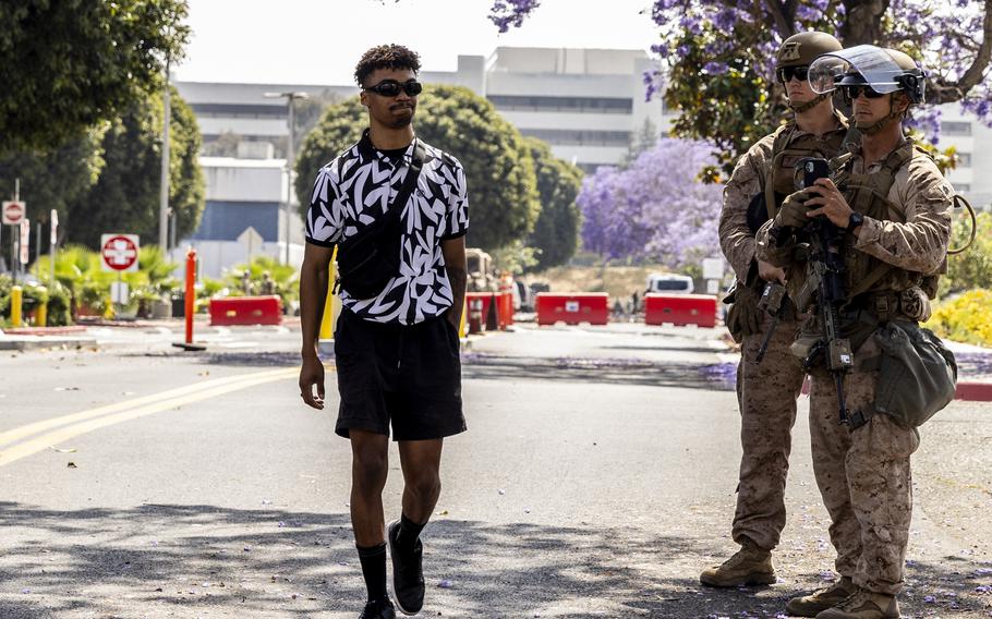 A man wearing a black and white floral shirt and sunglasses walks past two military service members in camouflage combat gear at a checkpoint on a street with a multi-story office building in the background.