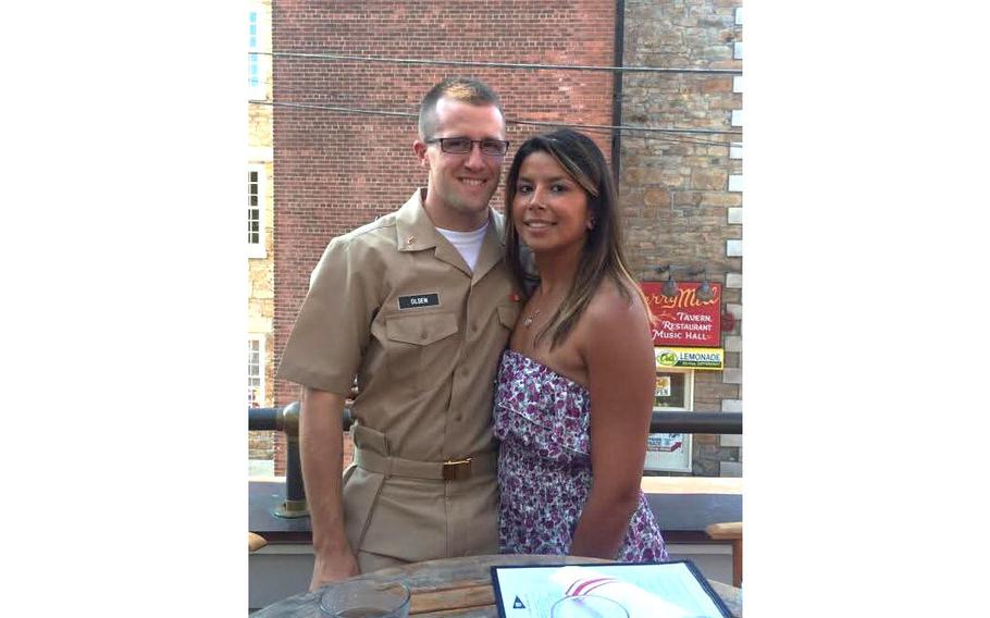 A man in a U.S. Navy officer service khaki uniform and glasses stands next to a woman in a strapless floral dress, both smiling on an outdoor patio with brick buildings in the background.
