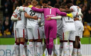 A soccer team wearing white uniforms with red accents is huddled together in a circle on the field, likely before the start of a match. The players have their arms around each other’s shoulders, showing unity and team spirit. One player, wearing a purple goalkeeper kit with the name “Kaiserslautern” on the back, stands out in the center. The background shows blurred spectators in the stands.