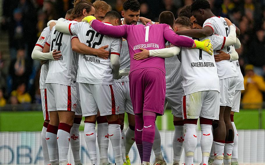 A soccer team in white kits huddles on the field, showing unity before a match. The goalkeeper in purple stands out at the center.
