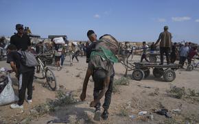 A man, seen from behind, carries the body of another man over his shoulder across sandy terrain.