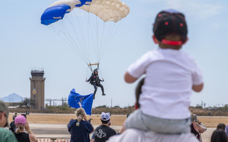 A young child sits on the shoulders of an adult and watches a person parachute from the air