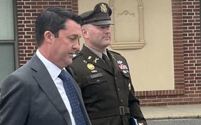An Army officer in dress uniform walks through a parking lot with a binder and book inhis left hand, while a man in a suit walks next to him.