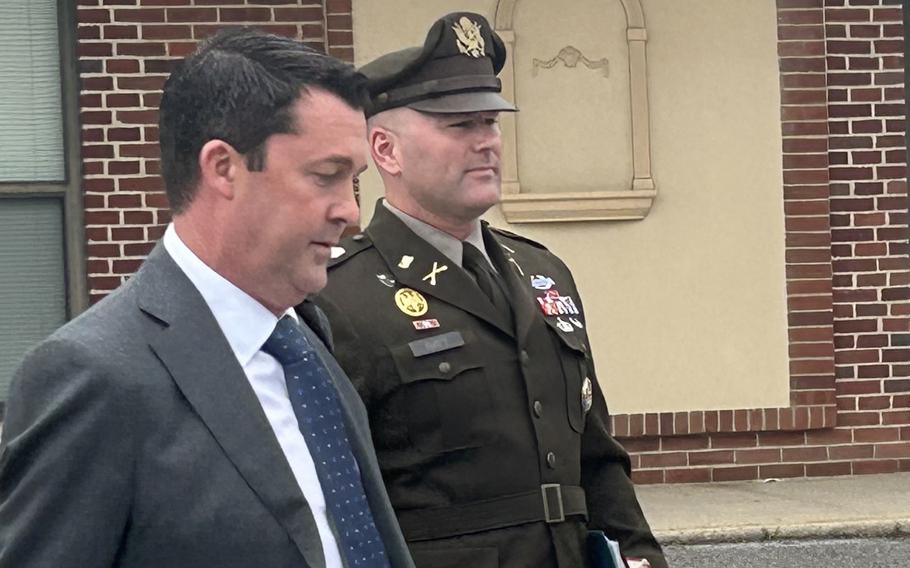 An Army officer in dress uniform walks through a parking lot with a binder and book inhis left hand, while a man in a suit walks next to him.
