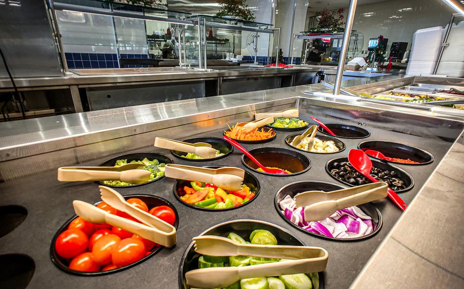 A close-up of a salad bar with tongs on top of the various salad components.