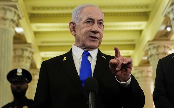 Benjamin Netanyahu, wearing a dark suit and blue tie, gestures with his hand as he speaks into a microphone while standing in a hallway in the U.S. Capitol building.