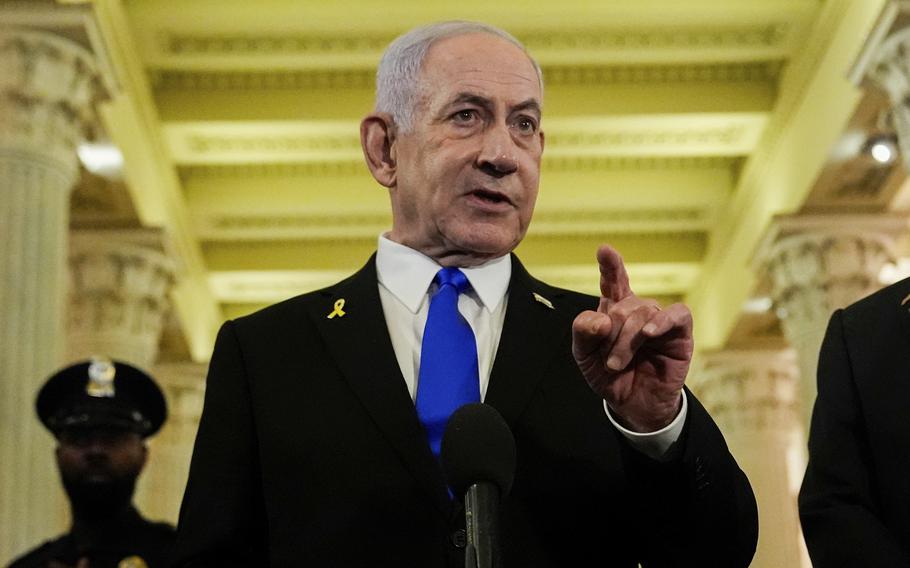 Benjamin Netanyahu, wearing a dark suit and blue tie, gestures with his hand as he speaks into a microphone while standing in a hallway in the U.S. Capitol building.