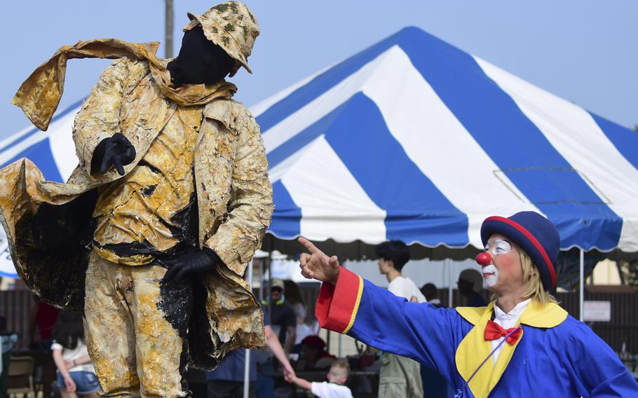A man dressed as a clown stands and points to a man on the left in camouflage uniform posing as a statue at an outdoor festival.