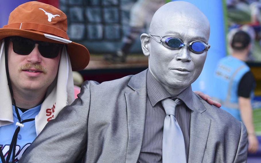 A man in an orange University of Texas hat poses next to a man dressed as a statue with a silver suit and tie and silver body paint.