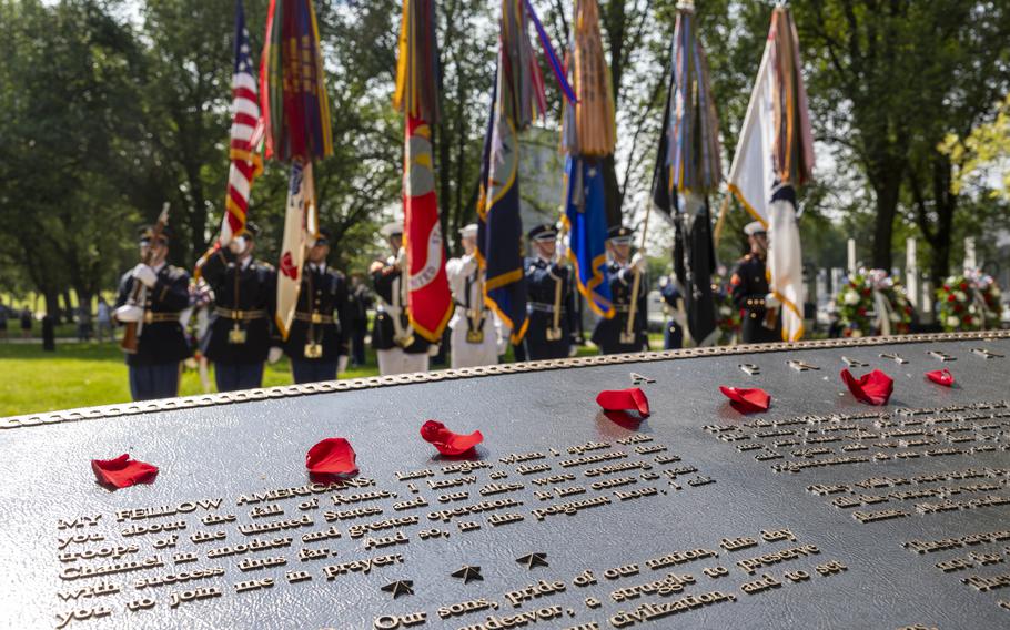 The text of an in-focus plaque in the foreground with a military honor guard and flags out-of-focus beyond it.