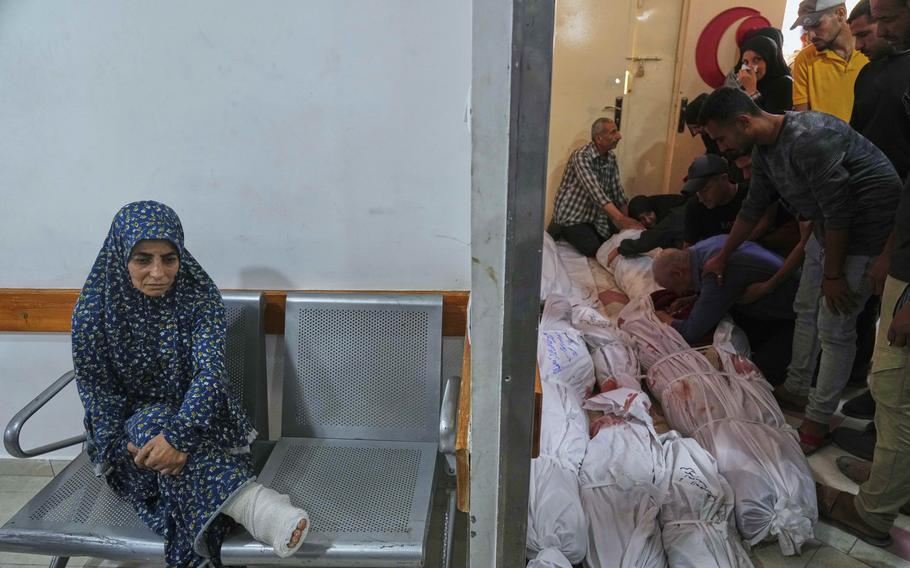 A woman sits outside of a room where men stack corpses killed in military strikes.