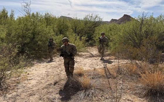 Three soldiers in combat gear and holding guns walk through desert shrubbery with the horizon in the background.