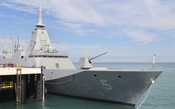A modern naval frigate with a distinctive angular, stealth design moored at a pier. The ship has a gray hull with the number “5” visible on its bow, along with a prominent pyramidal superstructure and various radar and communication arrays. A gun turret is visible on the forward deck. 