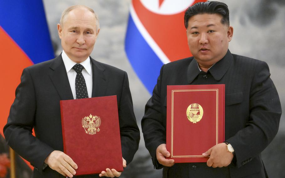 Vladimir Putin and Kim Jong Un pose next to each other while holding red folders with gold emblems in front of Russian and North Korean flags.