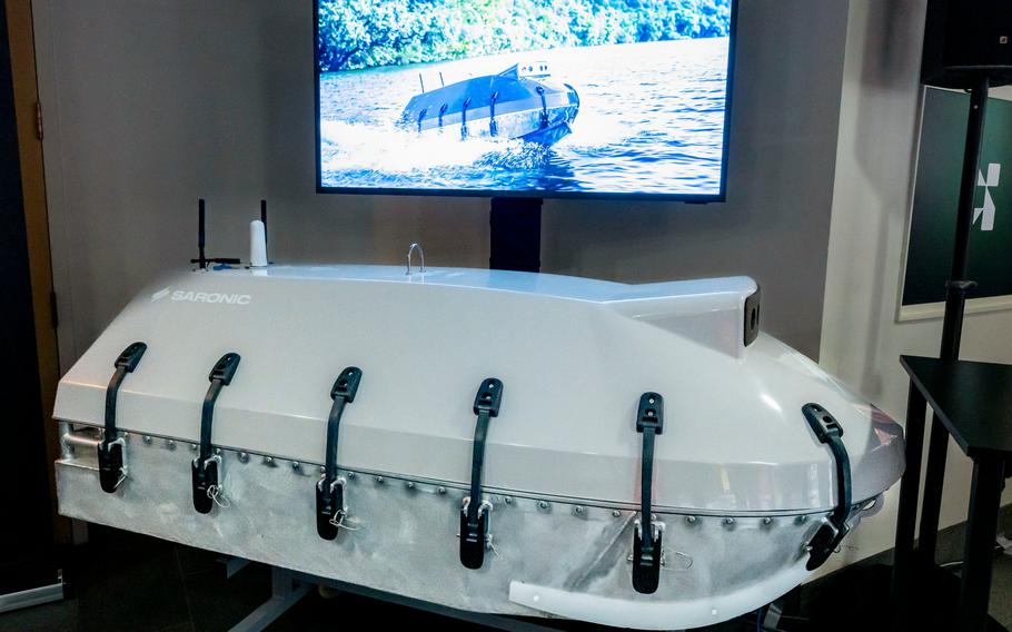 A white vessel seen indoors before a TV screen that shows the vessel at sea.