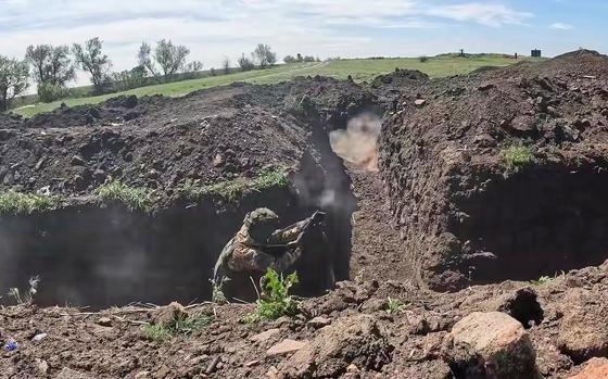A soldier in camouflage combat gear readies a weapon while standing in a trench dug into the ground.