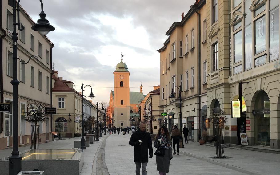 People walk down pedestrian 3 Maja, a shop-lined street in Rzeszow, Poland. Lit by a setting sun in the background is the Parish Church of St. Stanislaus and St. Wojciech.