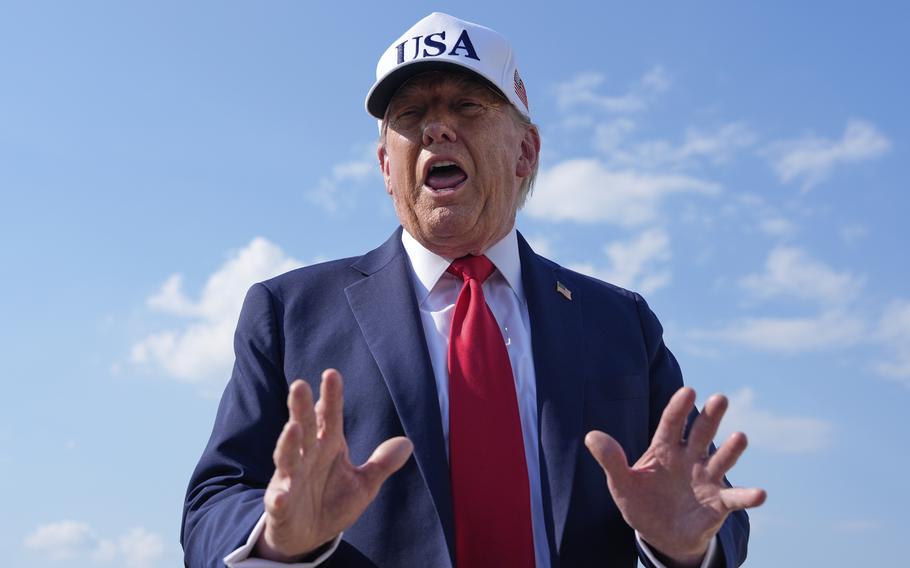 Donald Trump , wearing a dark suit and white hat and seen from a low angle, gestures with his hands as he speaks with blue sky behind him.
