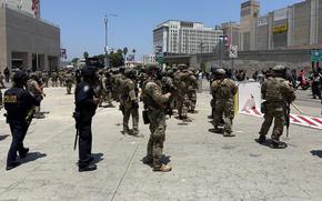 Troops in camouflage combat gear and holding weapons stand along a concrete walkway with police as protesters march by.