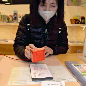 A Japanese woman wearing a facemask uses an orange stamp to make a mark on a piece of paper on the table in front of her.