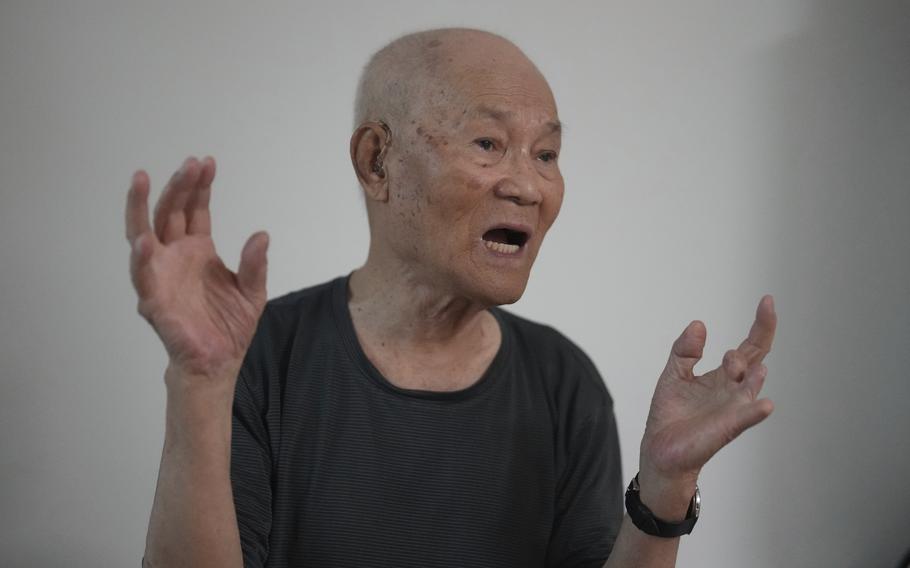 An elderly man in a black t-shirt speaks while gesturing with both hands.