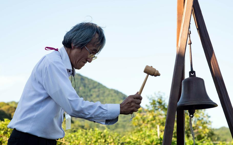 An elderly Japanese man wearing a white shirt and glasses uses a wooden mallet to ring a small bell hanging from a wooden frame, with a forested hill in the background.