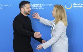 Volodymyr Zelenskyy reaches to embrace Giorgia Meloni against a blue poster or wall.