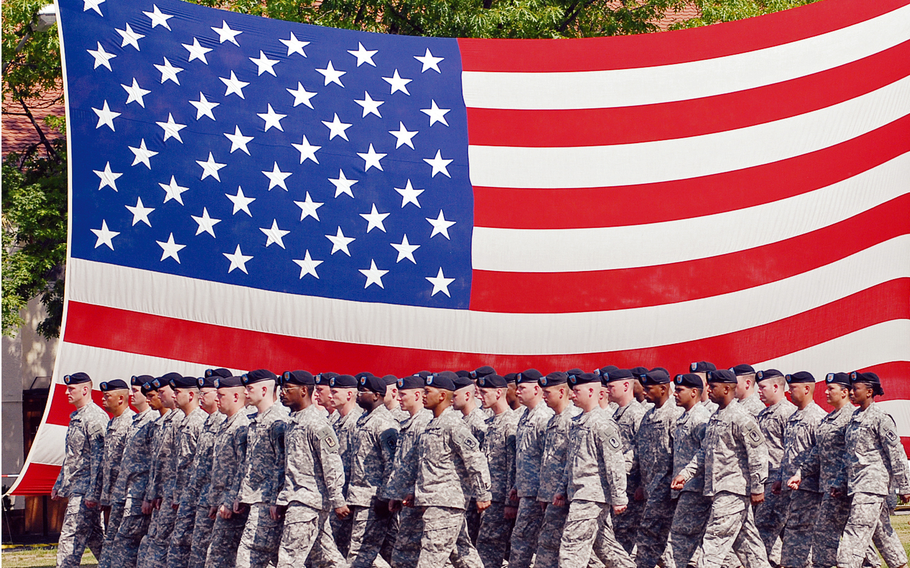 A formation of soldiers in camouflage uniforms and black berets marches in front of a large U.S. flag.