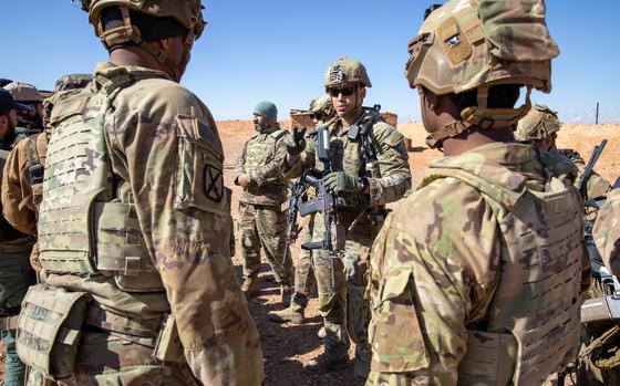 Several soldiers in camouflage combat gear and helmets stand together in a group with a desert skyline behind them.