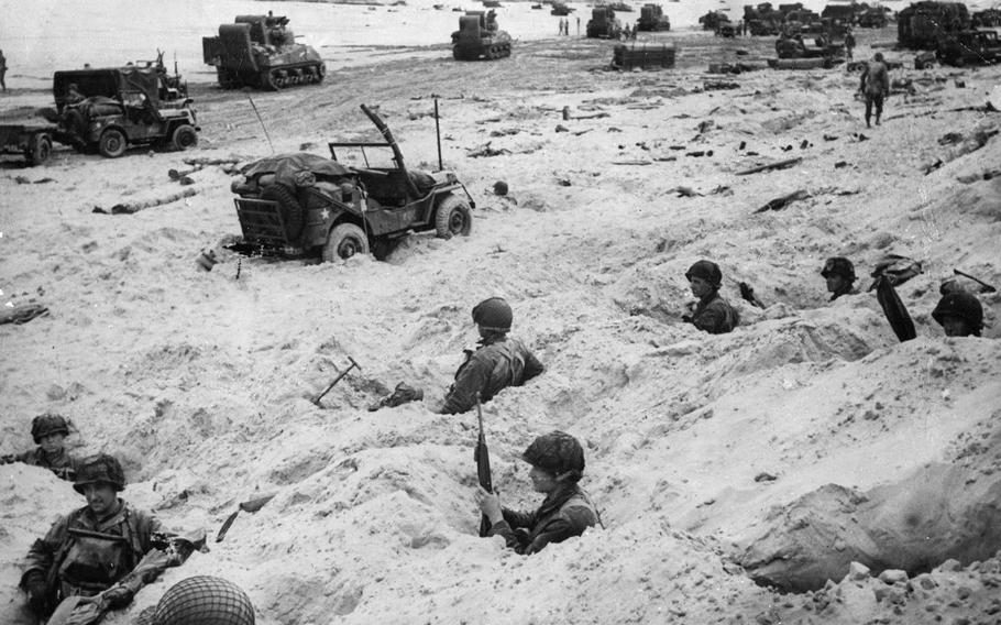 American soldiers — members of the Allied Expeditionary Forces which made the initial landings in France June 6, 1944 - keep watch from foxholes on the beachhead until it is cleared. In the background trucks, DUKWs, amphibious tanks and other equipment crowd the beach, while sea landing craft disgorge more troops and equipment. June 14, 1944
